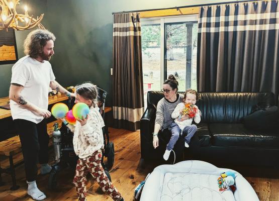 Woman holding child on couch while another girl walks with coloured balls in front of a man