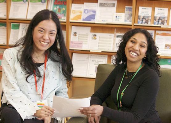 Two women with dark hair hold papers with shelves behind them