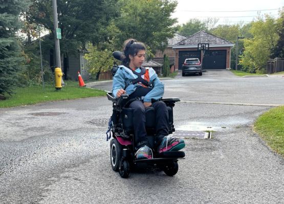 Teen with long dark hair drives power wheelchair in suburban neighbourhood.
