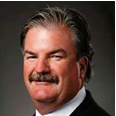 Man with brown hair and a moustache wearing a black blazer and striped tie