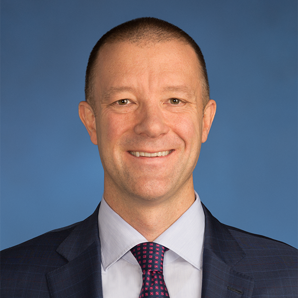 Man with brown, buzzed hair wearing a dark blue blazer, white shirt, and blue and red tie.