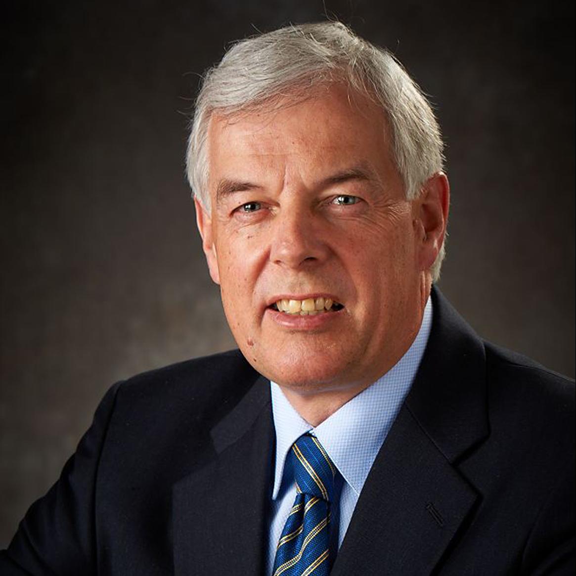 Man with gray hair wearing a dark blazer, light blue shirt, and a blue-striped tie