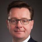 Man with short dark hair and black rectangular glasses. He's wearing a black blazer, white dress shirt, and a red and white striped tie