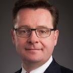 Man with short dark hair and black rectangular glasses. He's wearing a black blazer, white dress shirt, and a red and white striped tie