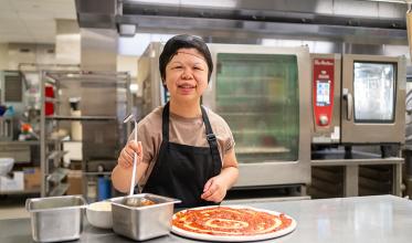 A smiling person in an industrial kitchen putting tomato sauce on pizza dough