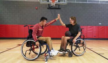 Two teenagers, both of whom use wheelchairs, high fiving in a gym