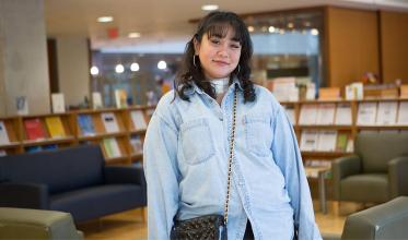 A young adult, who has a	 bandage on their neck, standing in a library, smiling