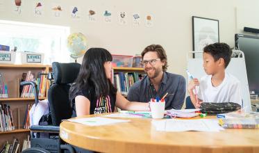 An adult sitting in the middle, to the left is a child on wheelchair and to the right is another child, in a library setting