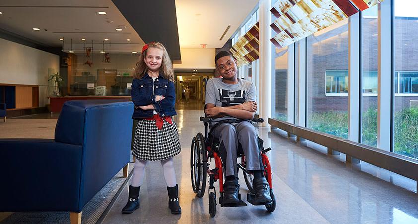 A child standing by the side of another child who is on a wheelchair