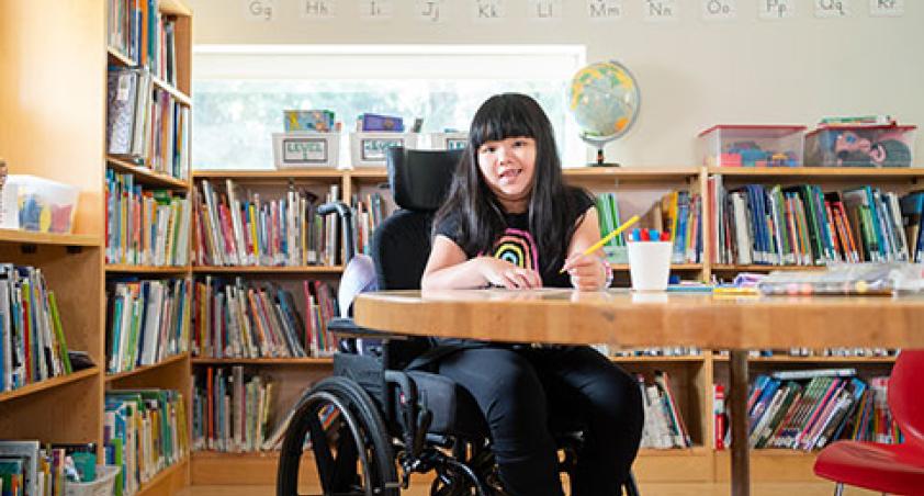 A child on a wheelchair doing some work on a desk, with a library setting at the back