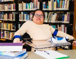 A young woman, who uses a wheelchair, sitting at a desk in front of some library bookshelves