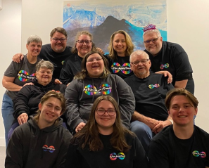 A smiling teen and their family sitting together for a family photo. Each family member is smiling and wearing black t-shirts and sweaters that say, "Autism Acceptance."