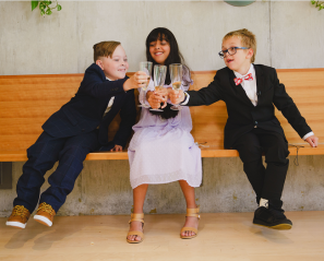Three children sitting on a brown indoor bench. The first child has light skin tone, brown hair and is wearing a blue suit. The second child has medium skin tone, long black hair and is wearing a white dress. The third child has light skin tone, short blonde hair and is wearing glasses and a black suit. The children are toasting their drinks.