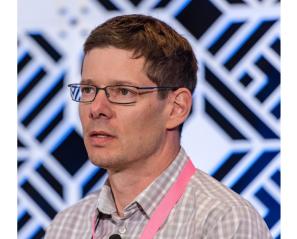 Dr. Jaimie Borisoff stands against a black and white checkered back drop wearing a light pink button up shirt, black glasses and short brown hair