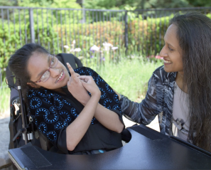 A young girl with medium skin tone and dark hair. She is smiling and sitting next to her mom outside. The young girl is using a wheelchair.