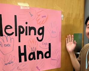 An adult volunteer holding the right hand in front of a hand draw poster with some hands on, and the words "Helping Hand"