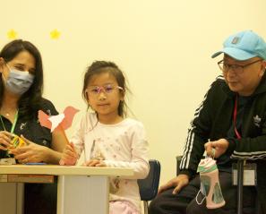 Girl at table holding pencil with pink paper bird on top wearing pink glasses with woman with dark hair on the left and man in a track suit jacket and a light blue baseball cap on the right