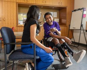 An adult sitting by the side of a teenager sitting on a wheelchair