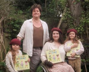 A man with brown hair stands behind a woman in a wheelchair and two girls who are each holding a children's book