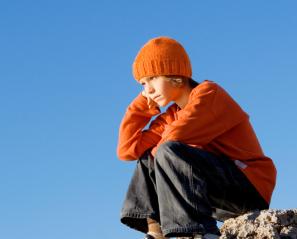 Child in orange sweater and hat sits on rock with head on fist