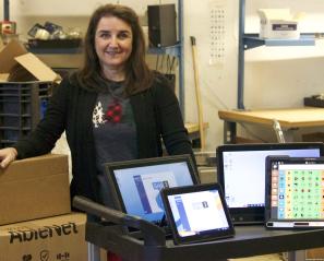 Woman with long hair stands in front of trolley with a number of computers displayed and boxes behind in warehouse