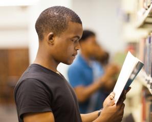 Young man in black t shirt with close cropped hair reads a publication in a library