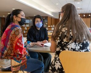 Three adults sit at a table together and have a conversation in the library.