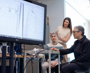 An adult sits and wears a brain computer interface headset while two other adults look at brain wave imaging.