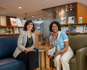 Two adults sit on arm chairs in the library, surrounded by shelves of books and pamphlets.