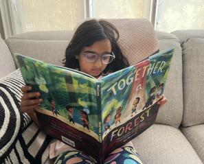 Girl with long dark hair and glasses reads large children's picture book