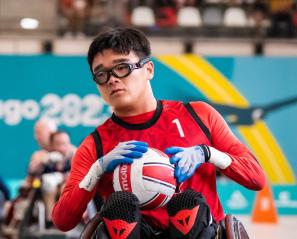 Man with dark hair and glasses wearing red athletic top and gloves holds ball in wheelchair