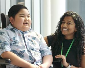 Boy with dark hair smiles in wheelchair beside woman with long dark hair who is also smiling