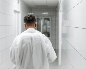 Back of man wearing white coat walking down hospital hallway.