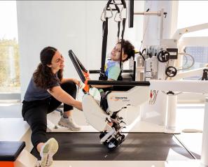 A child using a machine trying to learn how to walk, assisted by two hospital staffs