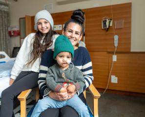 Two young children sit on their mom's lap, sitting on a chair in front of a hospital bed