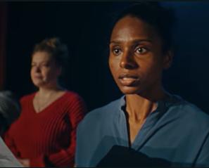 Woman in blue shirt stands in front of microphone while another woman is in the background