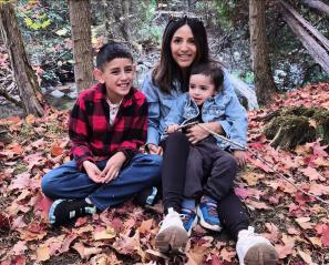 Two boys with dark hair sit in lap of woman with dark hair among colourful fall leaves