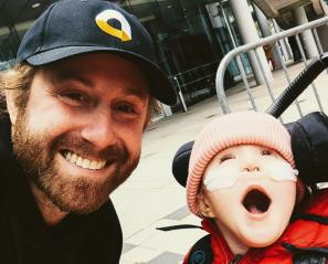 Man in baseball cap smiles beside young girl in stroller wearing nasal prongs