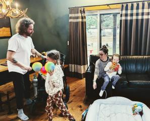 Woman holding child on couch while another girl walks with coloured balls in front of a man
