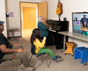 Child in yellow shirt sits in front of screen with icons with a woman on piano beside her and a man sitting behind her