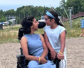 Teen girl in wheelchair and lavender outfit smiles at girl with bandana standing with arms on her chair