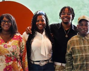 Photograph of four people standing side by side in front of a large orange circular backdrop with a green blurred background. They are wearing casual clothing, 