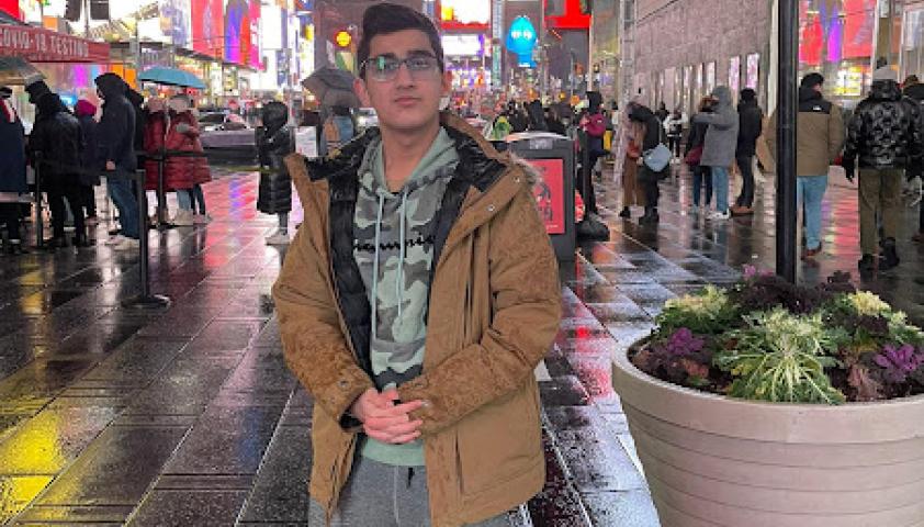 A teen with medium skin tone and black hair standing in a busy shopping centre outdoors. He is wearing a brown coat, grey sweatpants and red boots.