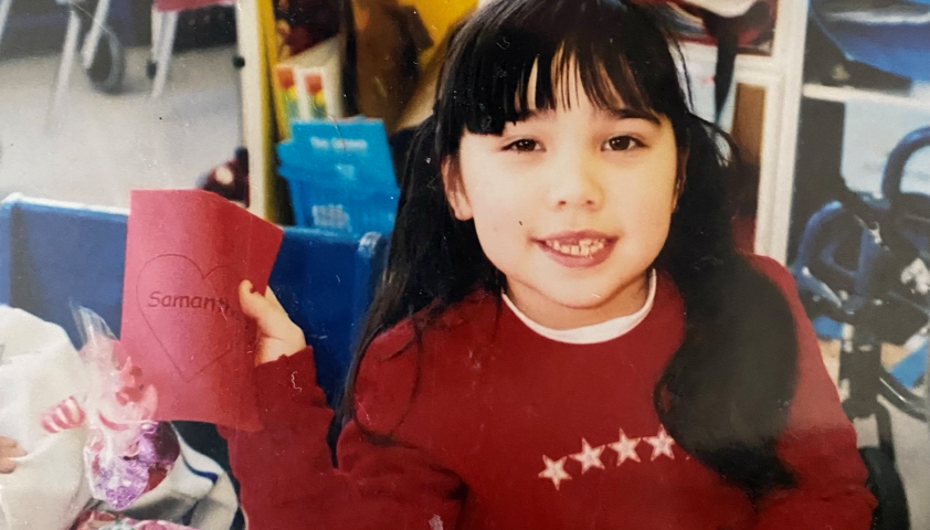 A young girl with light skin tone, wearing her long black hair in pig tails. She is wearing a red sweater and holding up a name tag that reads, "Samantha."