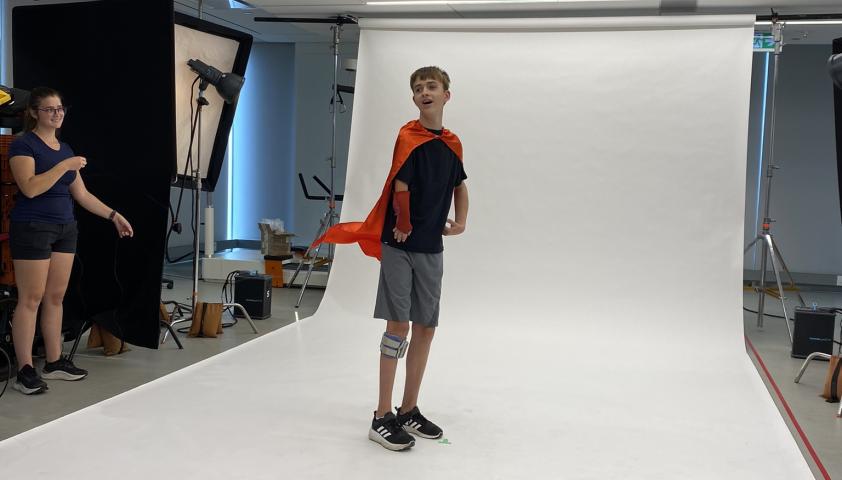 A young boy wearing a red cape stands in front of a white photo backdrop.