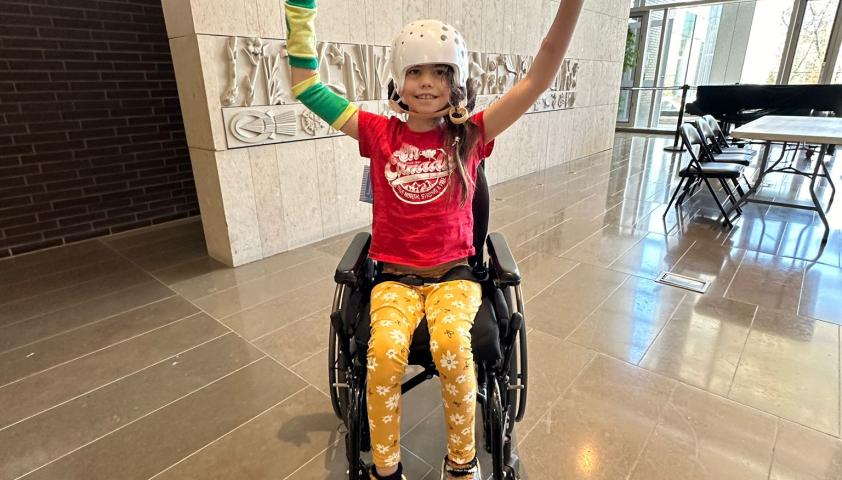 A child wearing a helmet and using a wheelchair raising her arms in excitement.