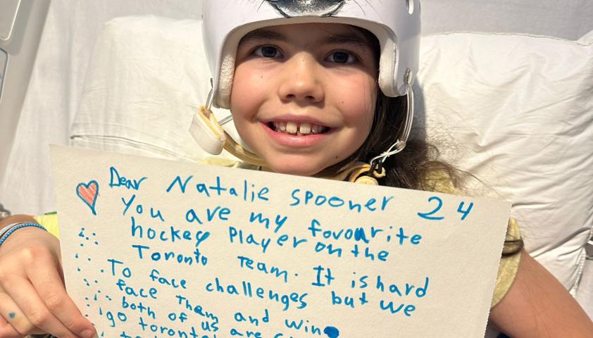 A child sitting in bed, wearing a helmet with a wolf painted on it and holding a sign.