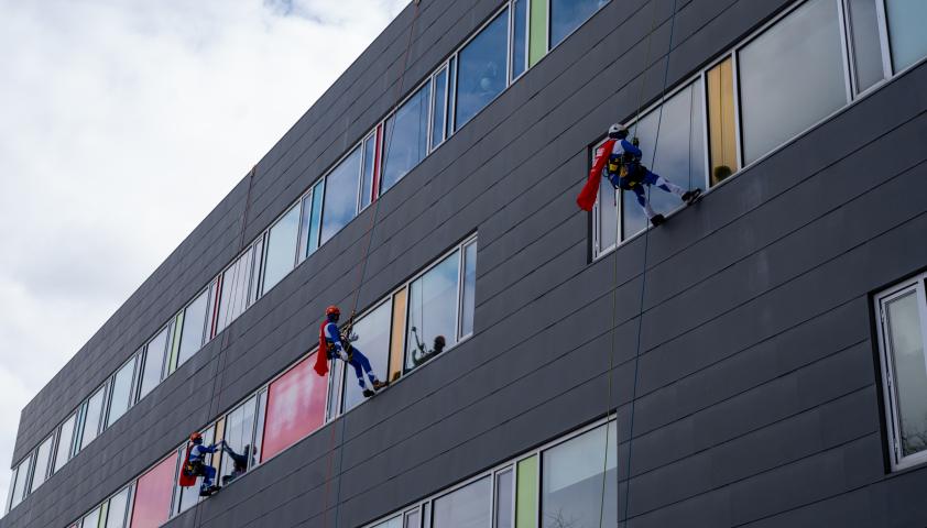 Three adults dressed as superheroes wear red capes and rapel down the exterior of the hospital.