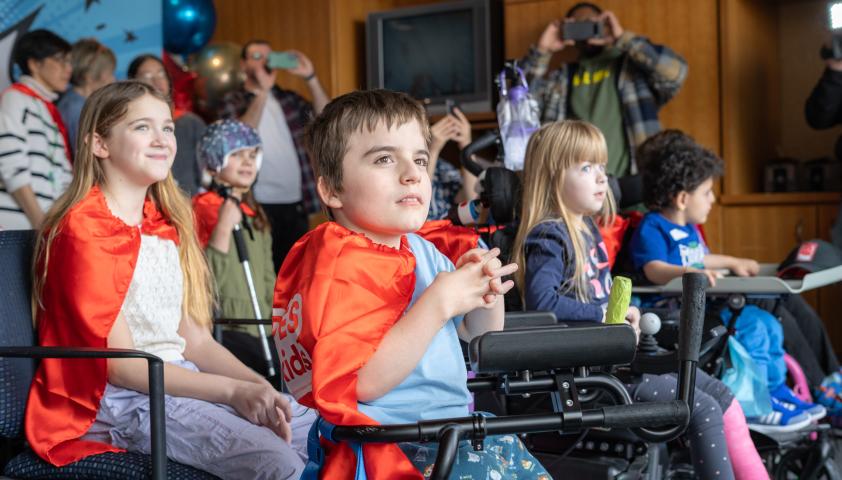 Children smile and wear red capes while looking out a window.