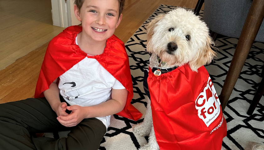 A young boy wearing a red cape sits with his dog who is also wearing a red cape.